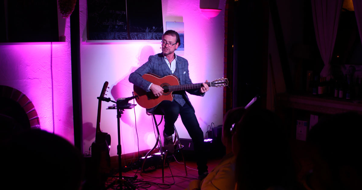 Mann sitzt auf einem Hocker und spielt Gitarre im Vordergrund des Bildes sitzen Menschen und schauen ihn an.