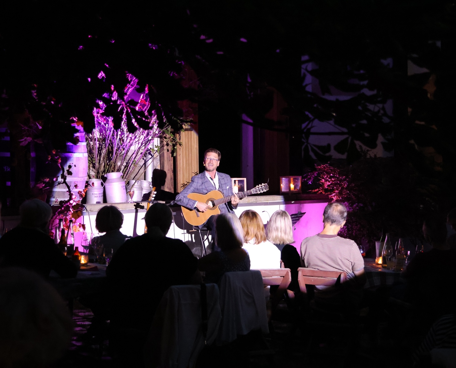 Mann sitzt draußen auf einem Hocker und spielt Gitarre. Er wird mit lila Licht beleuchtet und vor ihm sitzen Menschen und schauen ihn an. 