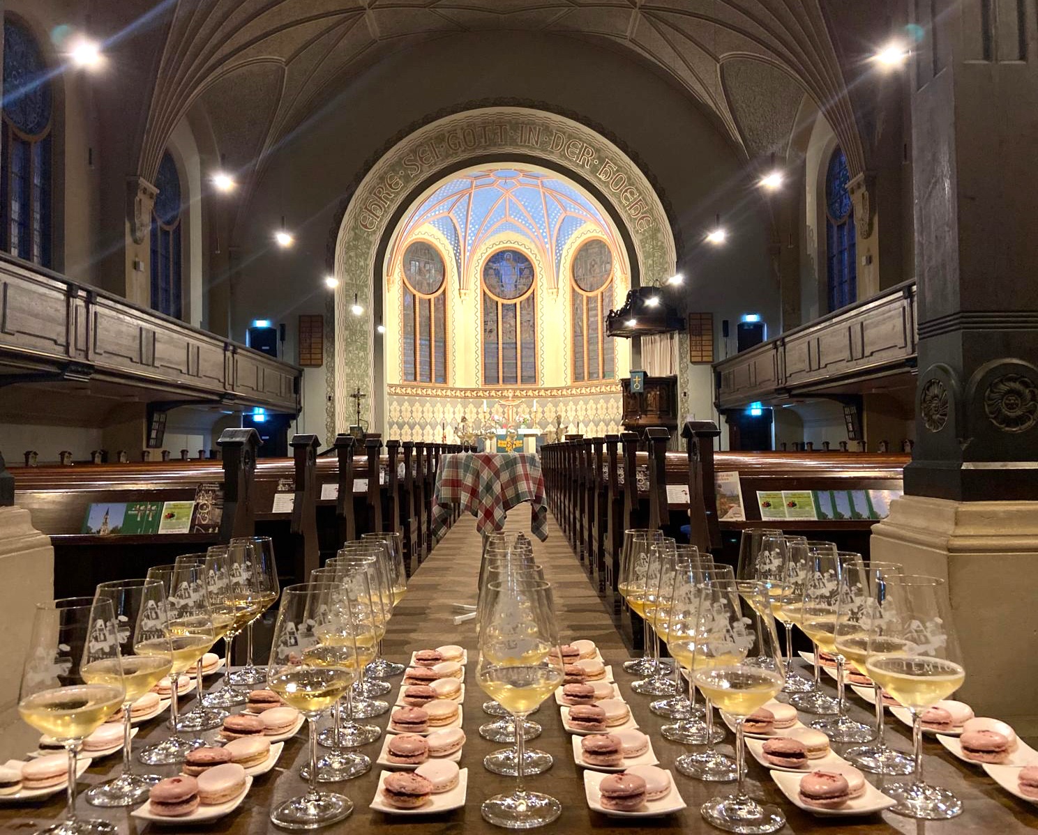 Im Vordergrund stehen fünf Reihen a sechs Weingläser mit Weißwein. Daneben jeweils ein  kleiner weißer Teller mit zwei rosa-farbigen Macarons. Im Hintergrund sind braune Kirchenbänke und mittig ist der hell erleuchtete Altarraum.