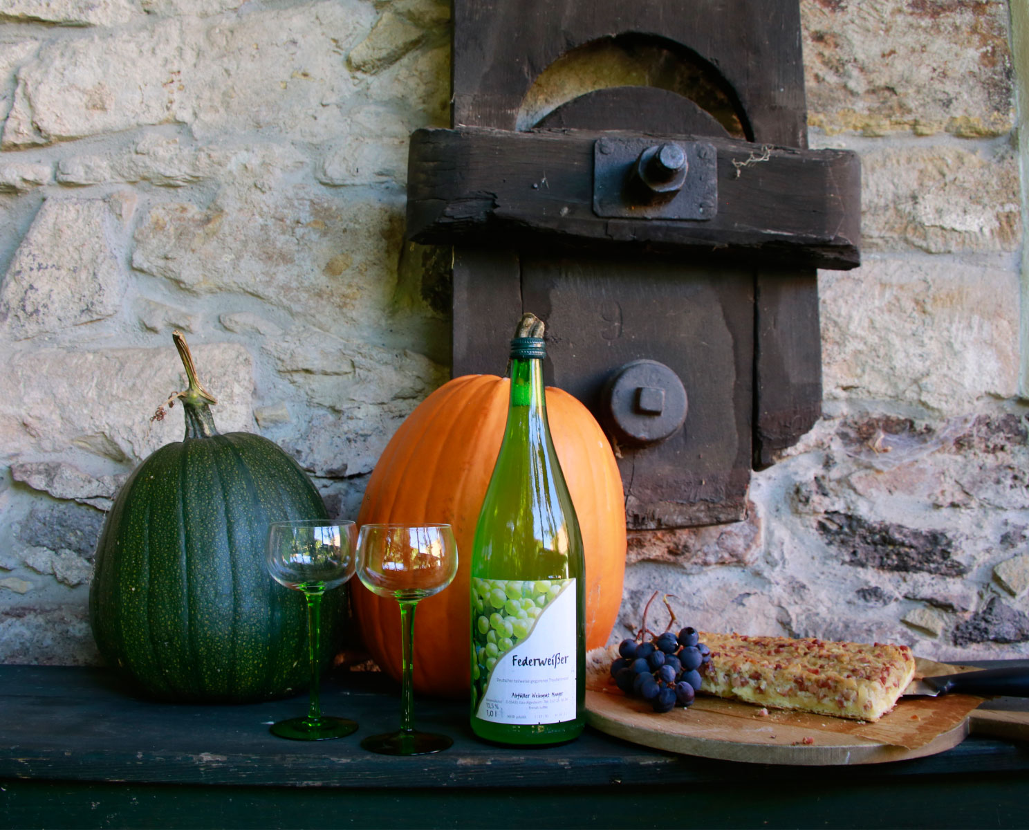 Zwei Kürbisse auf einer Bank, davor stehen zwei Gläser und eine Flasche. Auf einem Brett liegt ein Stück herzhafter Kuchen und Trauben in einer herbstlichen Szenerie.